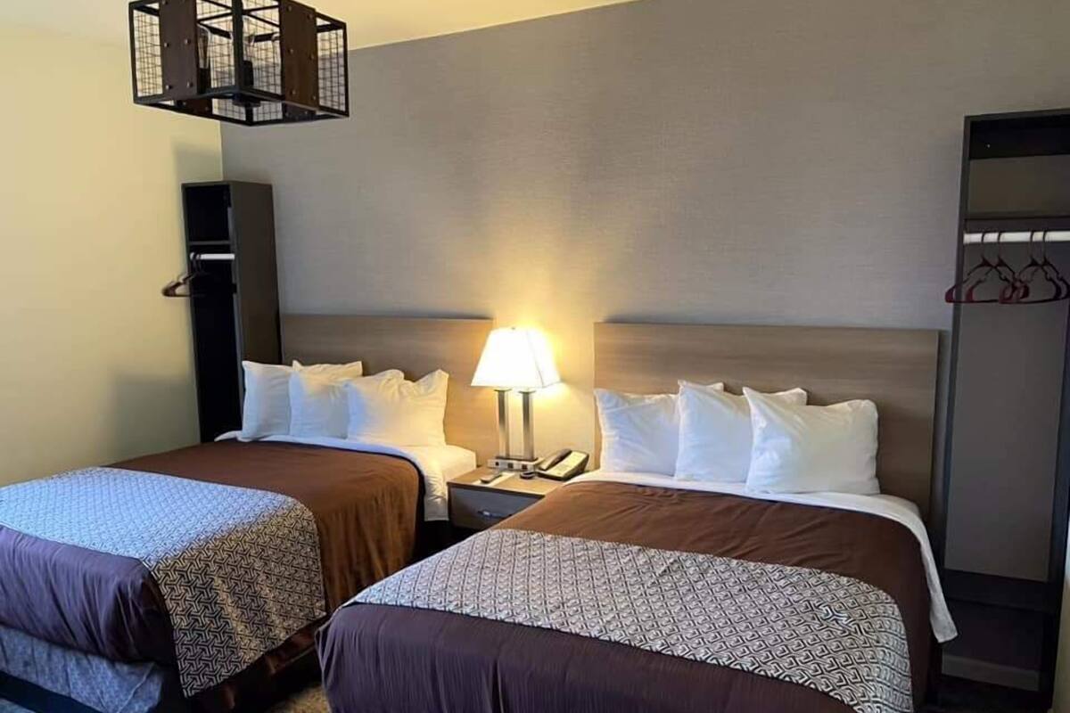 Quarto de hotel com duas camas, mesa de cabeceira com abajur, guarda-roupa e luminária pendente.