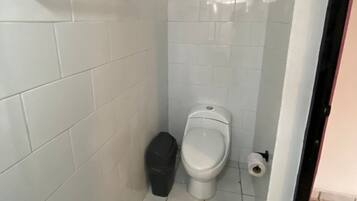 A small, white-tiled bathroom with a toilet, a trash bin, and a toilet paper holder.