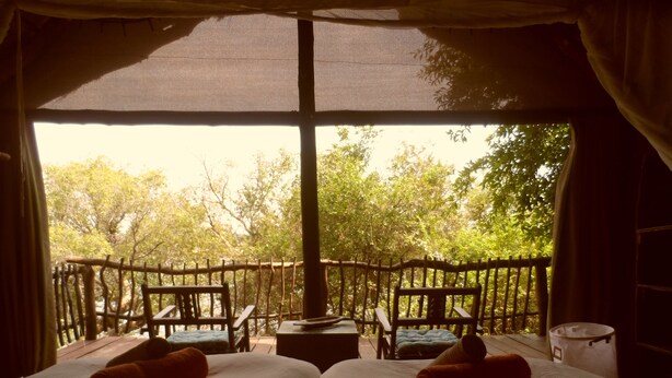 A room with two beds, a small table, and a chair, with a view of trees through a large window.