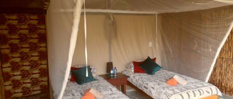 Een kamer met twee bedden, elk met een hemelbed en beddengoed met een patroon. Er staat een klein tafeltje met een waterfles en een kussen tussen de bedden. De kamer heeft een rieten dak en muren met een patroon.