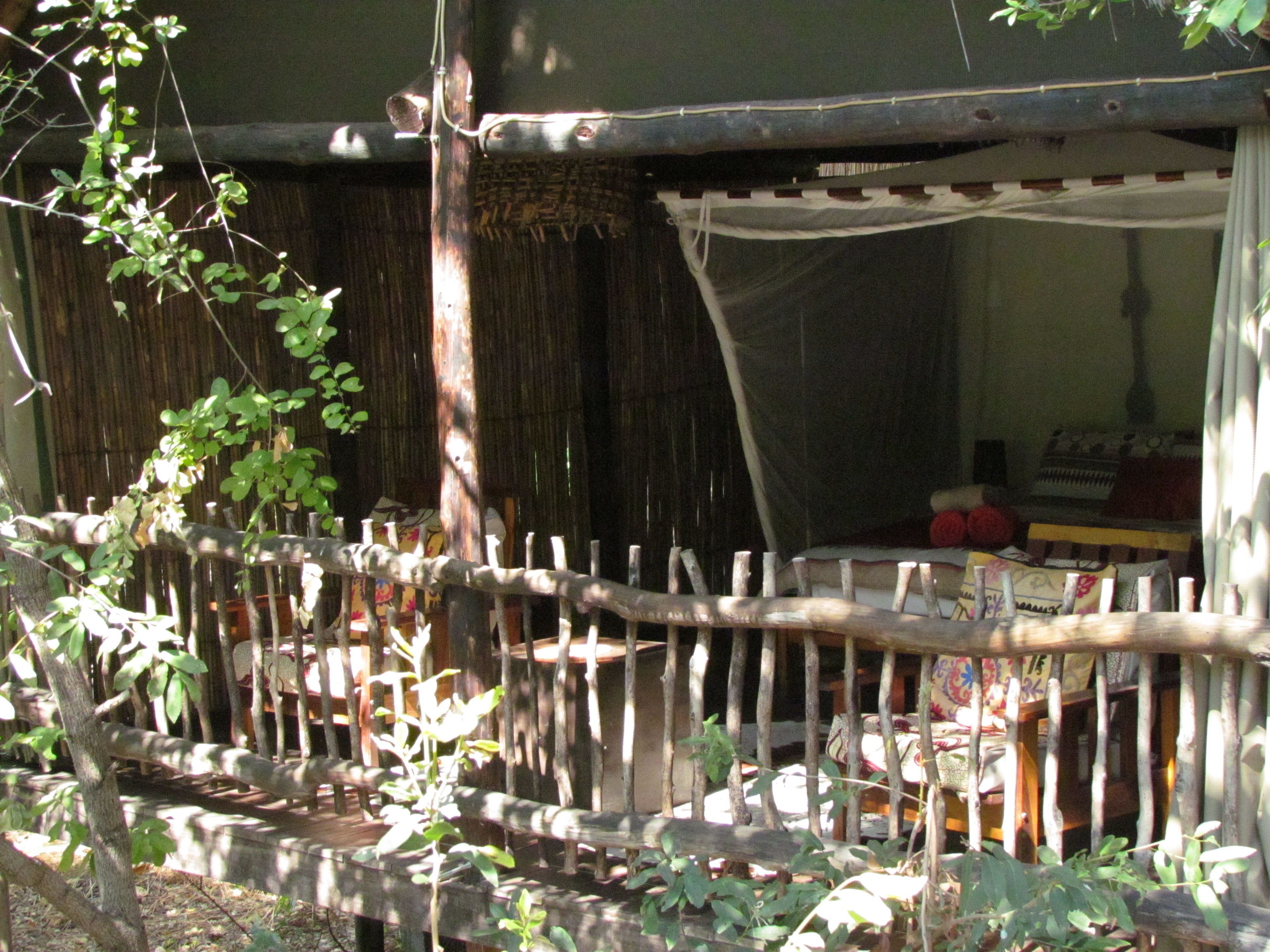 Uma cerca de madeira rústica com toldo tecido, um banco de madeira e uma pequena mesa com livros e um objeto vermelho.