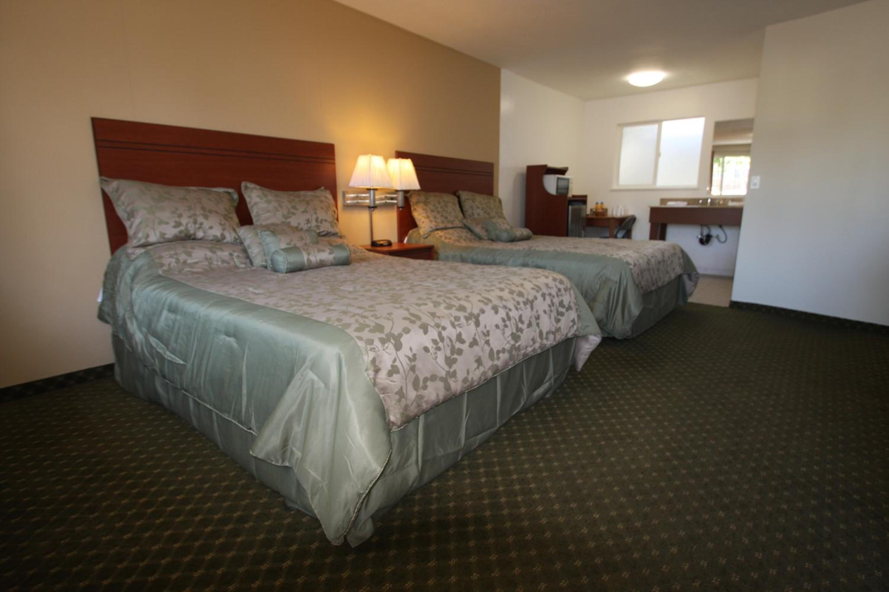Une chambre d’hôtel avec deux lits, une table de chevet avec une lampe, une petite table avec un miroir et une salle de bain avec un lavabo et un miroir.