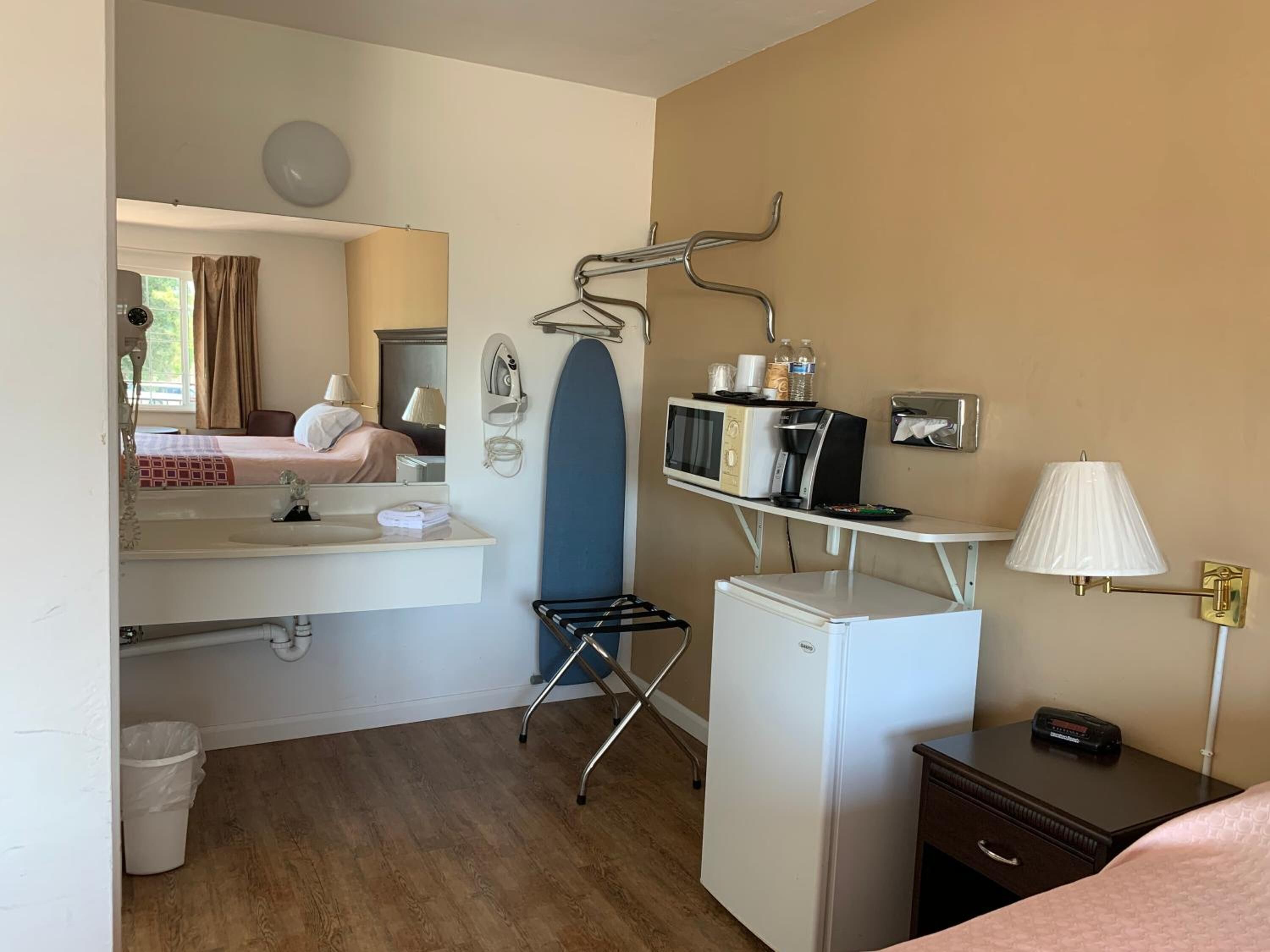 A compact hotel room with a kitchenette, a small refrigerator, a microwave, an ironing board, and a bedside table with a lamp.