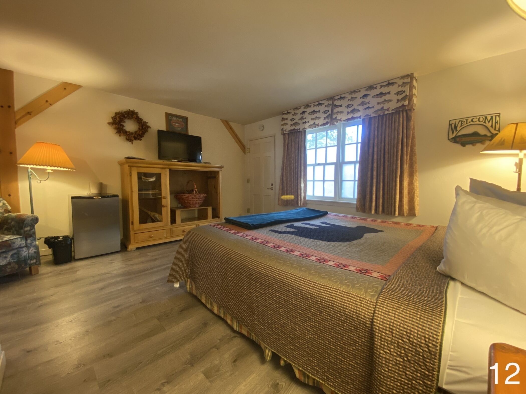 A cozy bedroom with a bed, a television on a cabinet, a window with curtains, and a welcome sign.