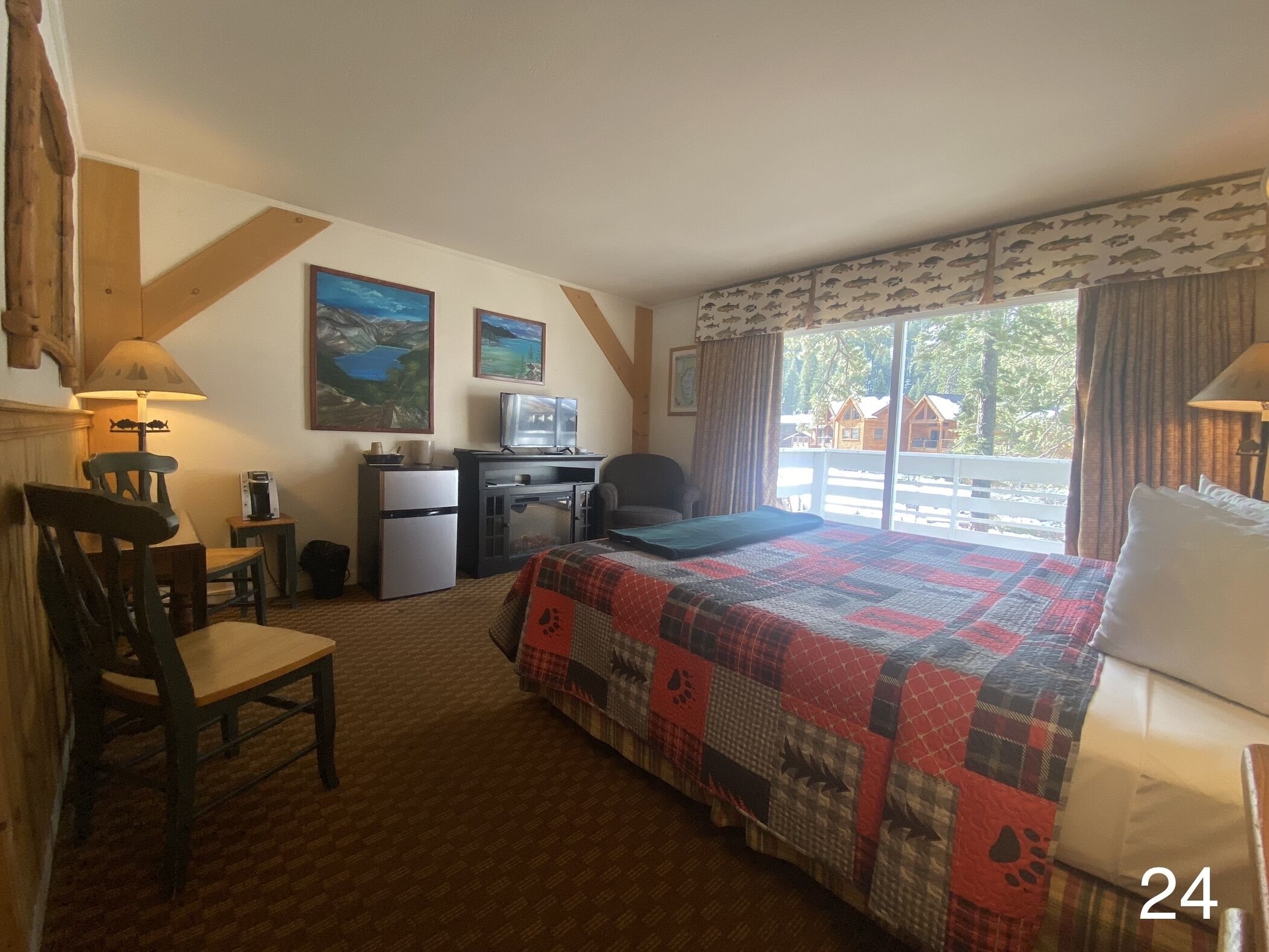 A hotel room with a bed, desk, chair, and a TV. There are two paintings on the wall, a window with a view of the outside, and a balcony with a railing.