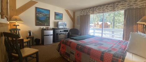 A hotel room with a bed, desk, chair, and a TV. There are two paintings on the wall, a window with a view of the outside, and a balcony with a railing.