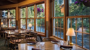 A restaurant with wooden tables and chairs, large windows with a view of a river and greenery, and hanging purple flowers. - River Ranch Lodge & Restaurant (Olympic Valley)