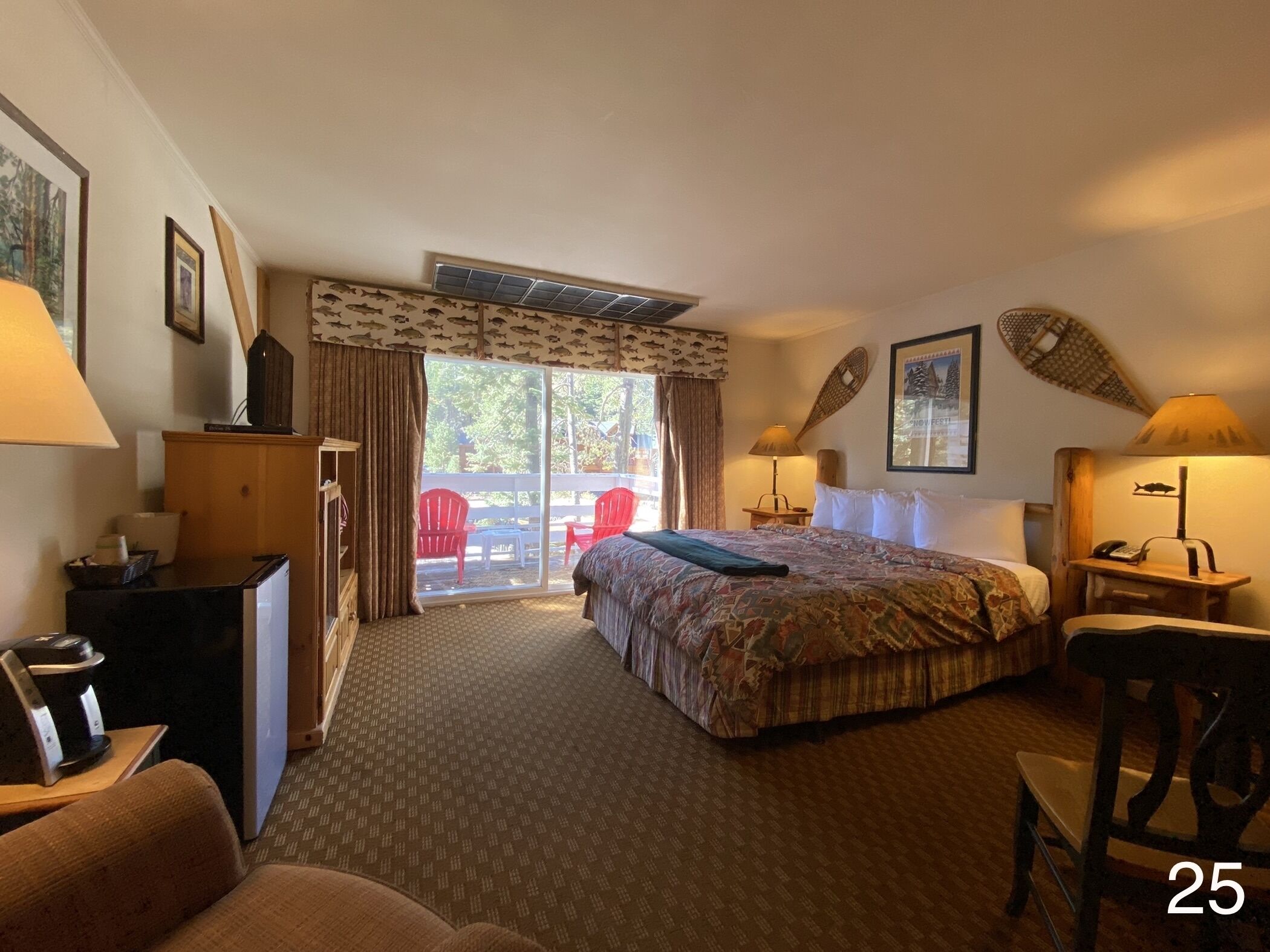 A hotel room with a bed, a desk, a chair, a dresser, and a sliding glass door leading to an outdoor area with red chairs.