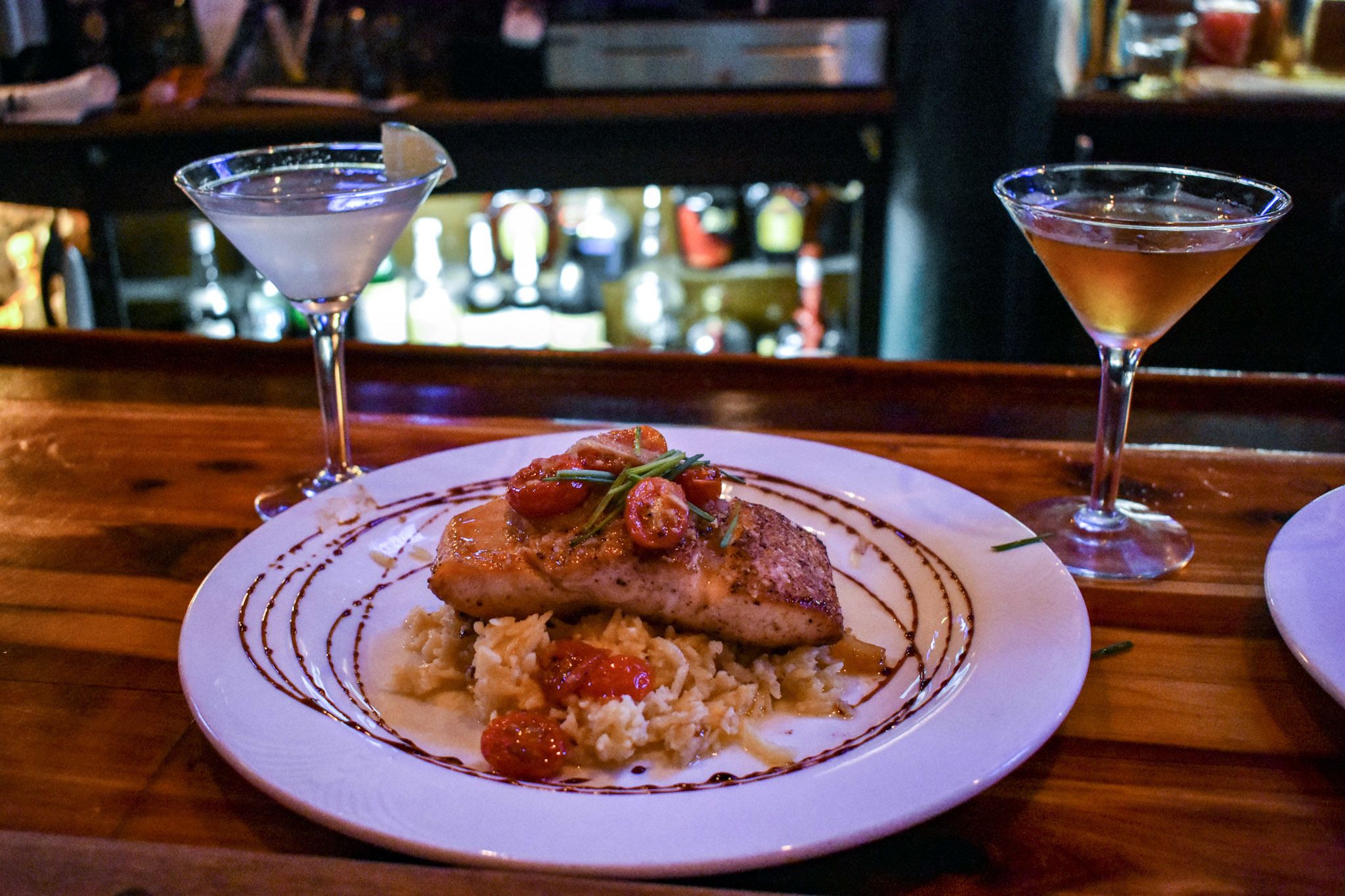 Un plato de comida con un filete de pescado, tomates cherry y una guarnición, acompañado de una copa de martini.