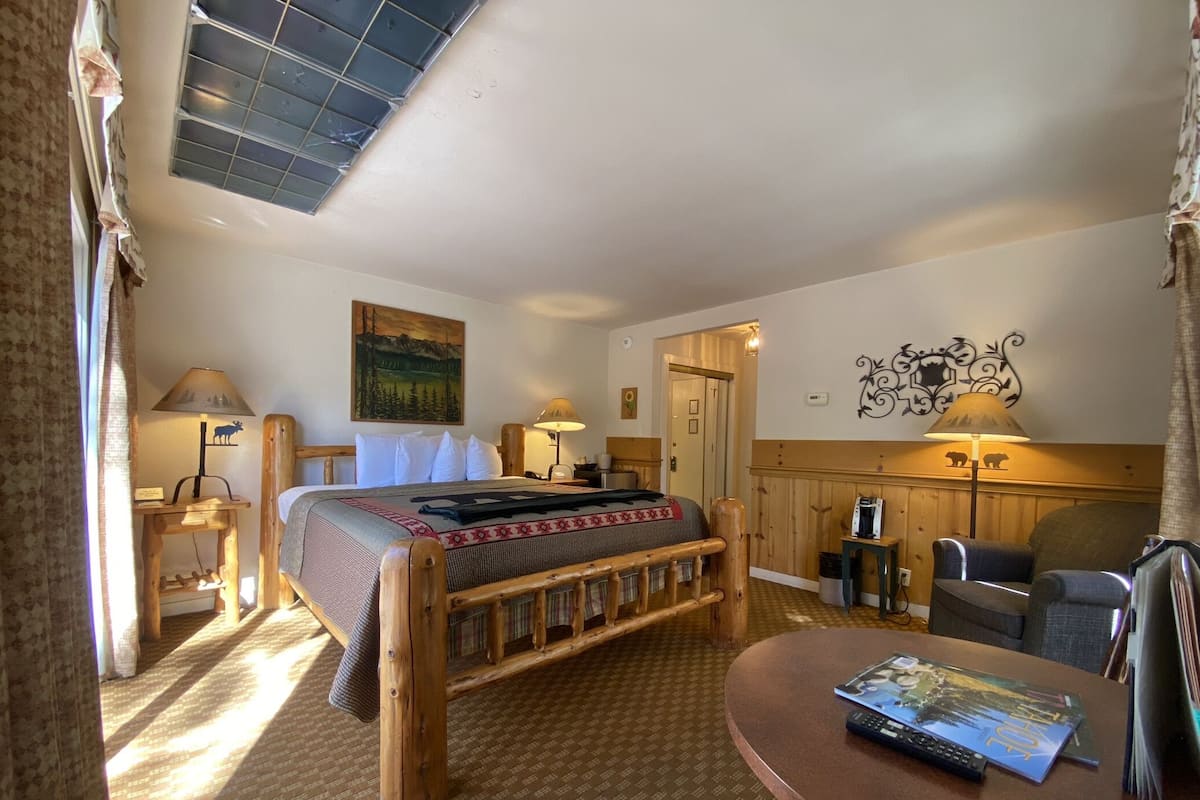 A cozy room with a wooden bed, a desk, and a chair. There's a painting on the wall and a lamp on the desk.
