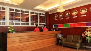 A reception area with a wooden reception desk, two receptionists, a sofa, a chandelier, and clocks on the wall. - Hotel K Yangon (Yangon)
