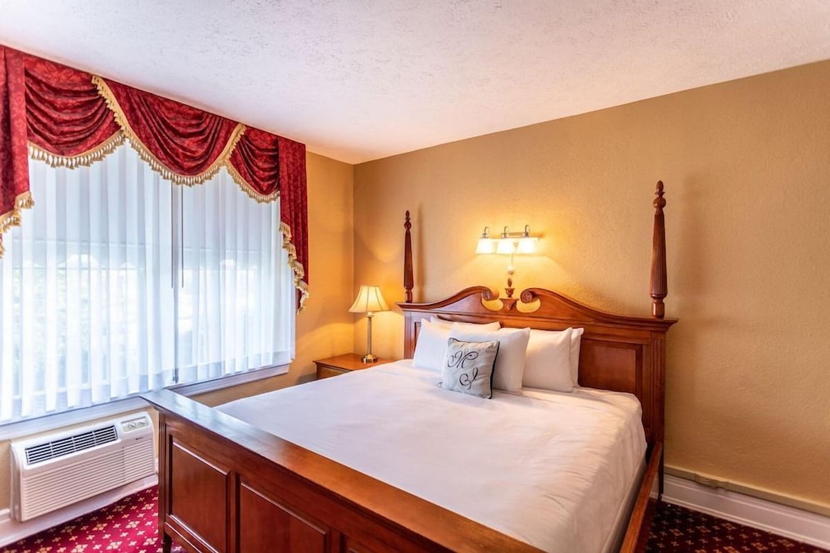 A hotel room with a wooden bed, a nightstand with a lamp, a window with curtains, and a wall-mounted light fixture.