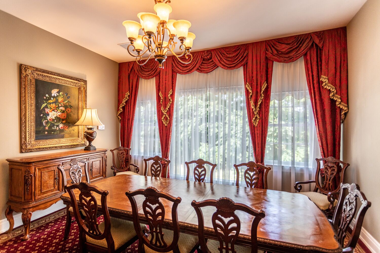 A dining room with a wooden table and chairs, red curtains, a chandelier, and a framed painting on the wall.