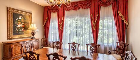 A dining room with a wooden table and chairs, red curtains, a chandelier, and a framed painting on the wall.