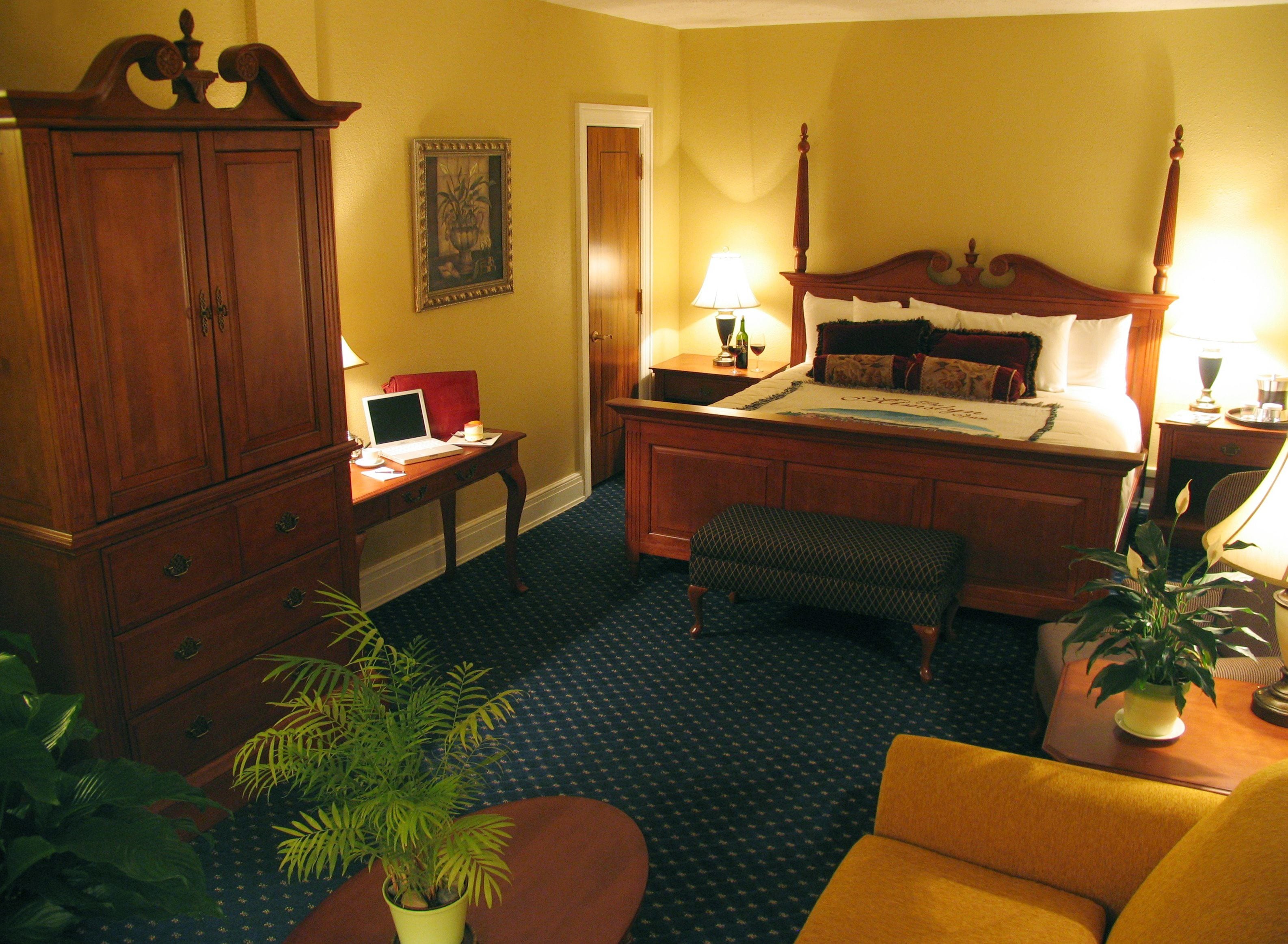 A hotel room with a large wooden bed, a desk with a laptop, a chair, a small table with a plant, and a large wooden cabinet.