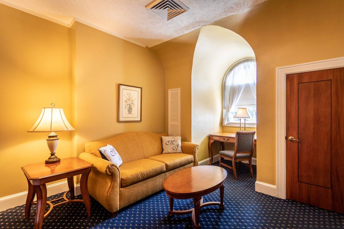A cozy room with a sofa, a small table, a chair, and a desk. There is a door and a window with curtains.