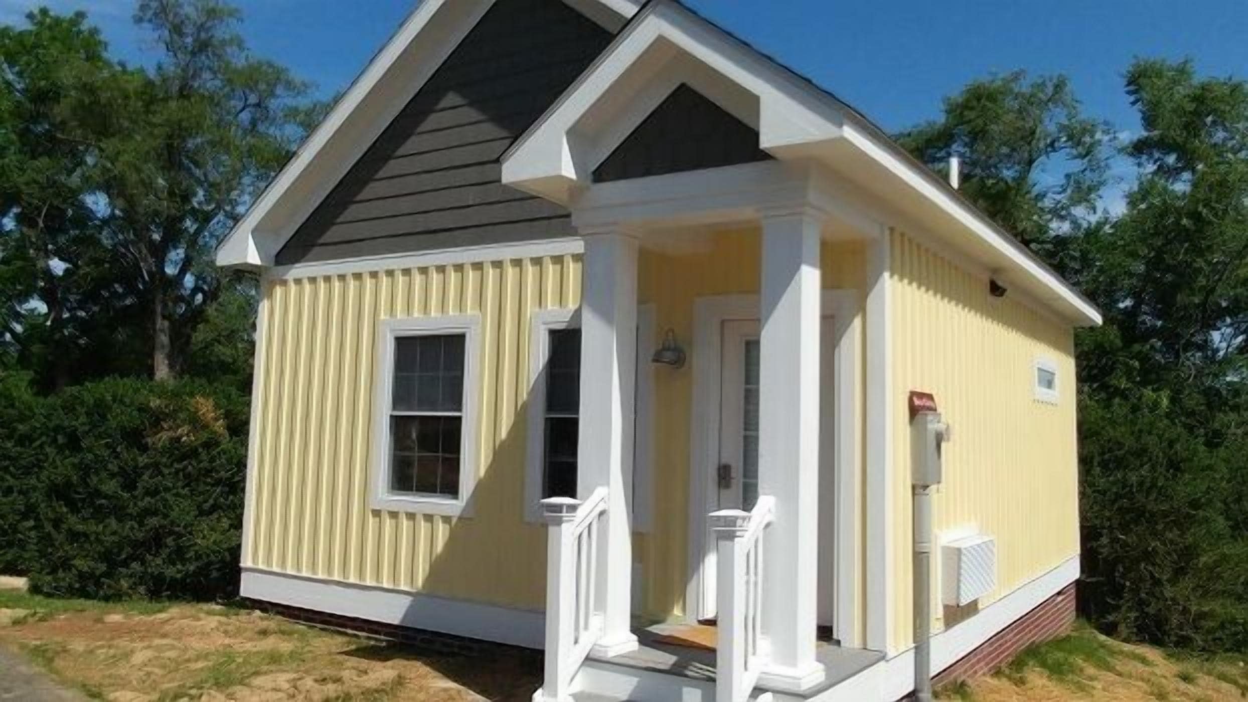 A small, single-story house with a gabled roof, a front porch with white columns, and a small window on the side.