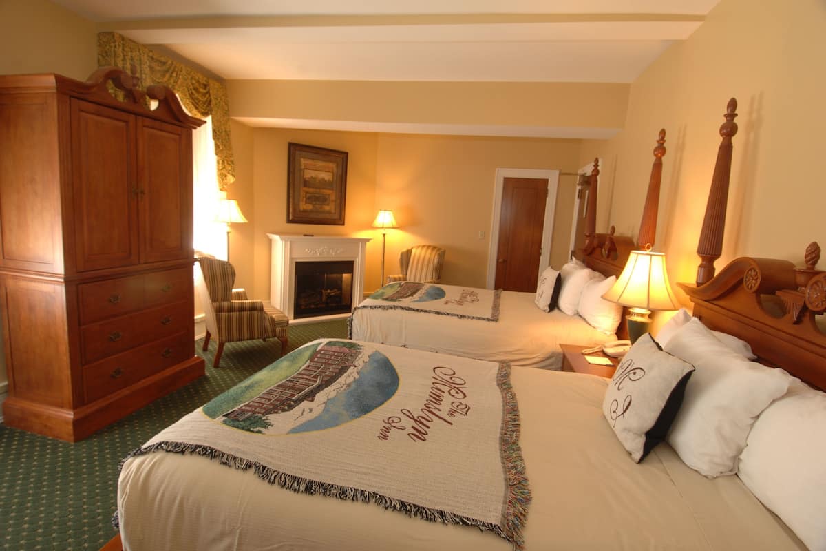 A hotel room with two beds, a wooden wardrobe, a chair, a fireplace, and a lamp.