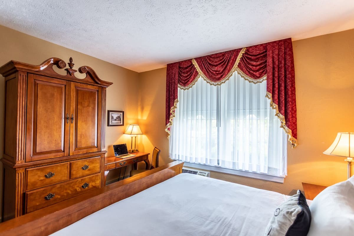 A hotel room with a large bed, a wooden wardrobe, a desk with a laptop, and a window with red curtains.
