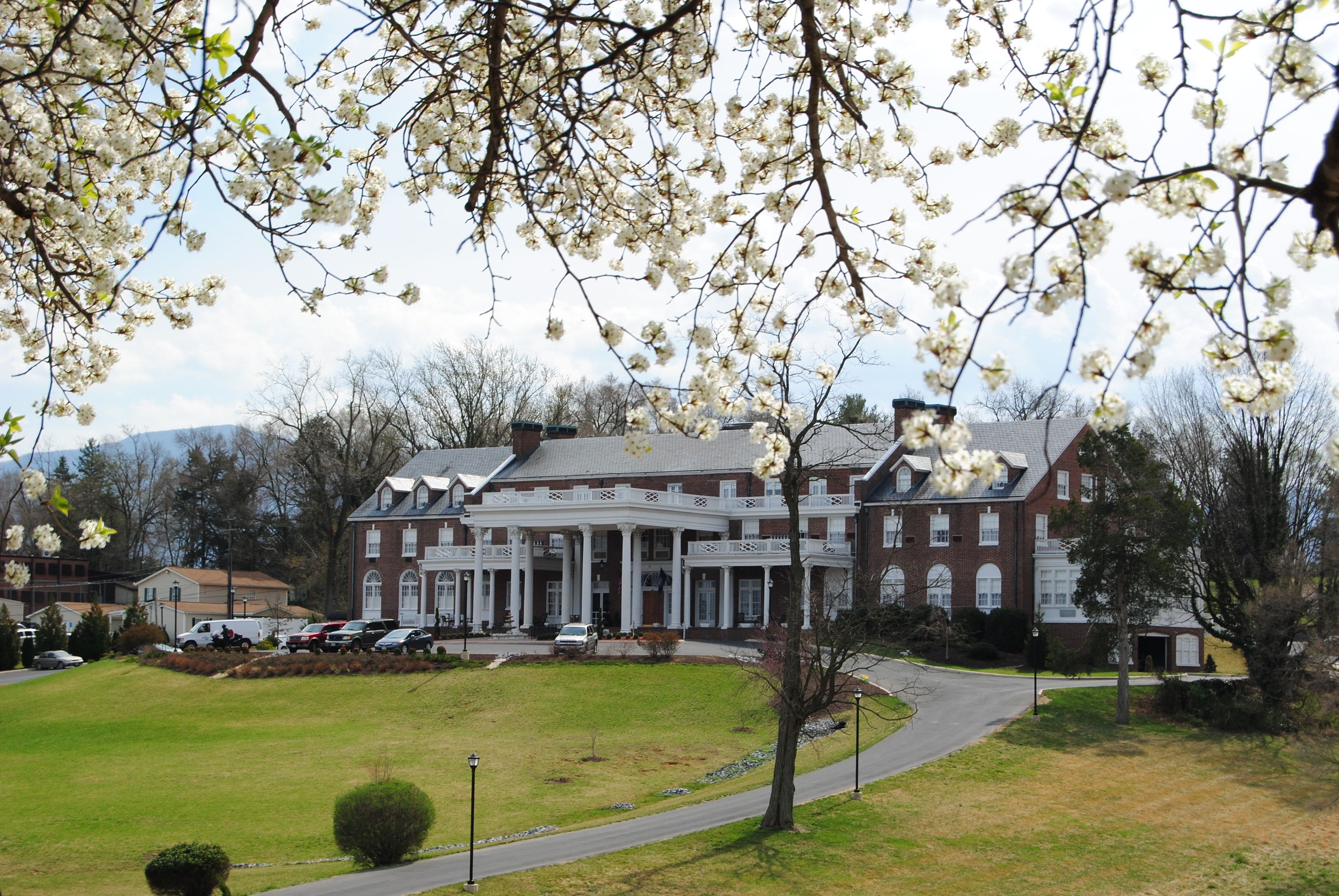 A large brick building with a central portico and multiple windows, surrounded by a well-maintained lawn and a parking area.