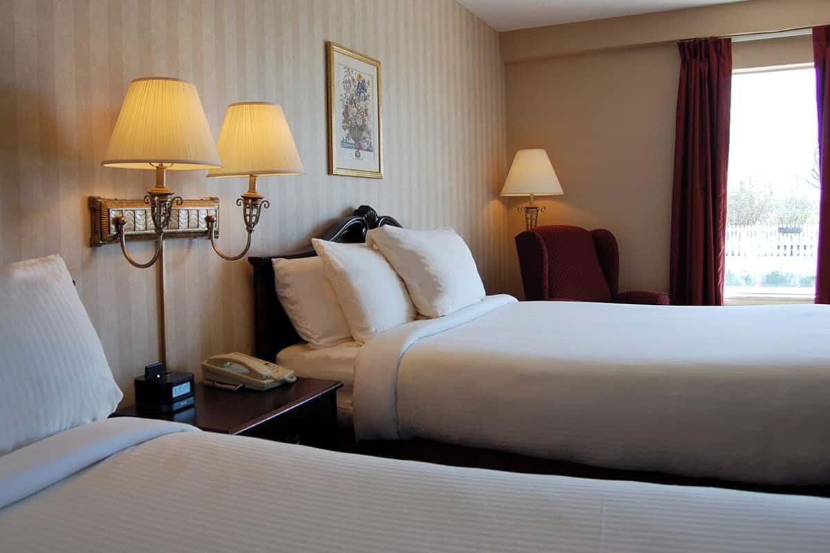 A hotel room with two beds, a nightstand with a phone, two wall-mounted lamps, and a framed picture on the wall.