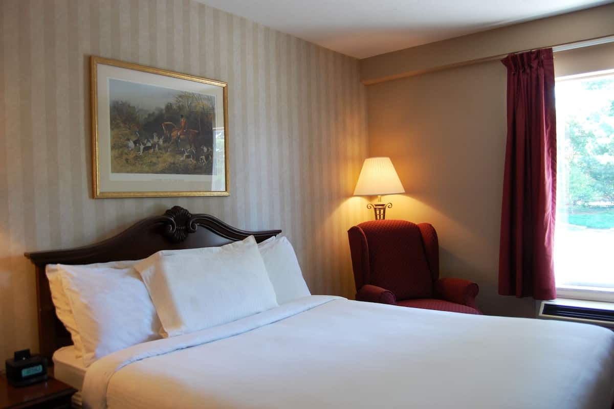 A hotel room with a large bed, a nightstand with a clock, a framed picture on the wall, a red armchair, a lamp, and a window with curtains.