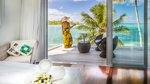 A room with a large window overlooking a tropical beach, a bed with a white cover, and a wicker chair. - Crystal Blue Lagoon Villas (Rarotonga)