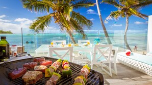A beachside dining area with a grill, tables, chairs, and a view of the ocean. - Crystal Blue Lagoon Villas (Rarotonga)