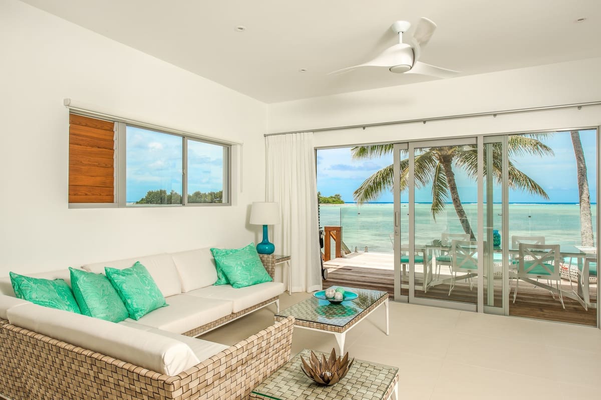 A modern living room with a white sofa, green cushions, a wicker coffee table, and a view of the ocean through large glass doors.