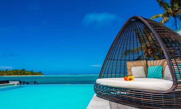 A wicker infinity pool with a clear blue ocean view, a lounge chair with cushions, and a palm tree. - Crystal Blue Lagoon Villas (Rarotonga)