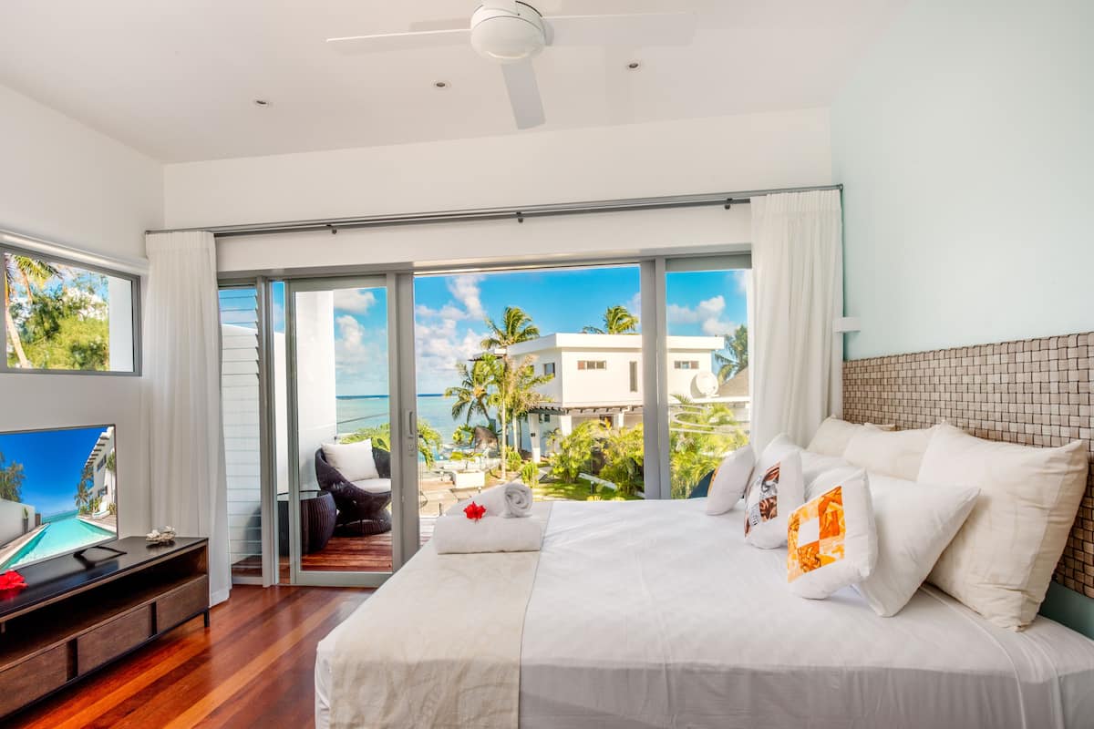 A modern hotel room with a large bed, a TV, and a balcony with a view of the ocean and palm trees.