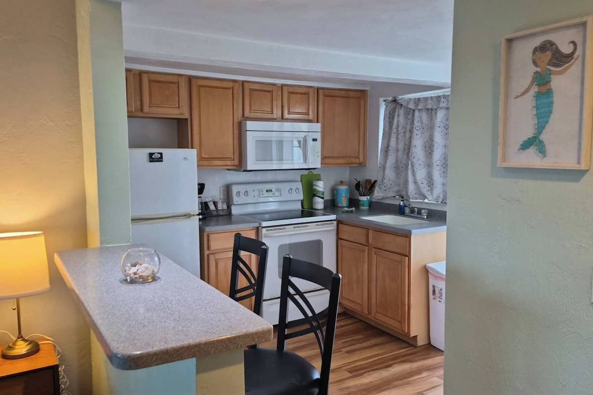 A compact kitchen with wooden cabinets, a white refrigerator, a microwave, a stove, and a sink. There is a small dining table with two chairs.