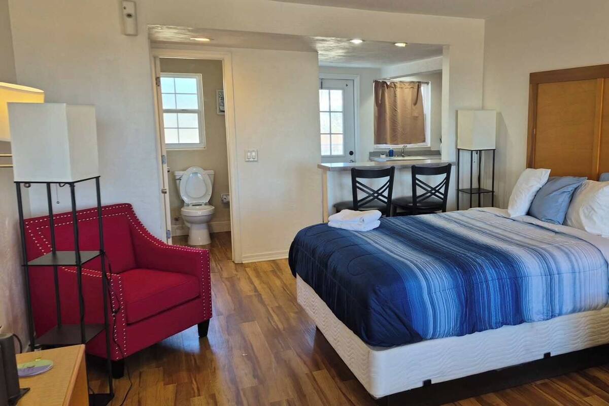 A hotel room with a large bed, a red armchair, a gray armchair, a small table, and a bathroom visible through an open door.