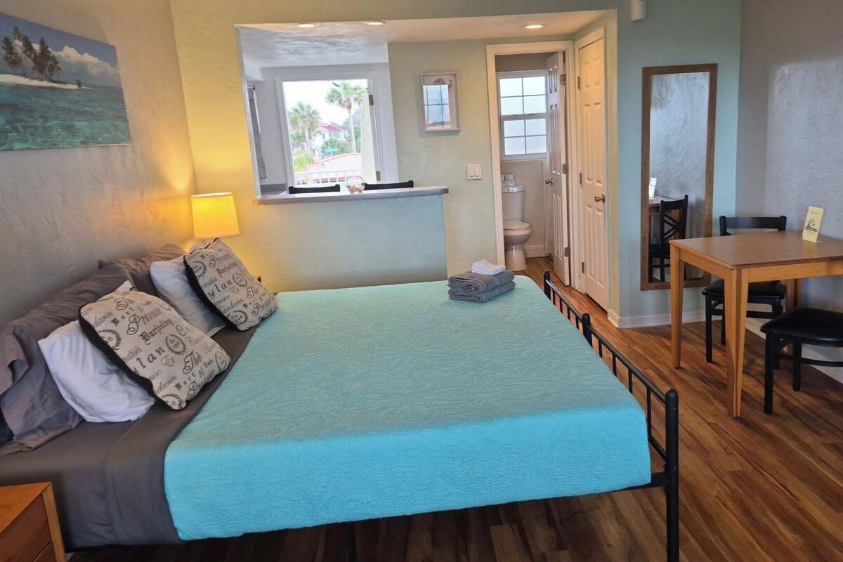 A bedroom with a bed, a bedside table, a lamp, a window, a door, and a view of the outside.