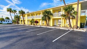 A yellow motel with a parking lot, palm trees, and a white car parked outside. - Studio 1 Motel (Daytona Beach)