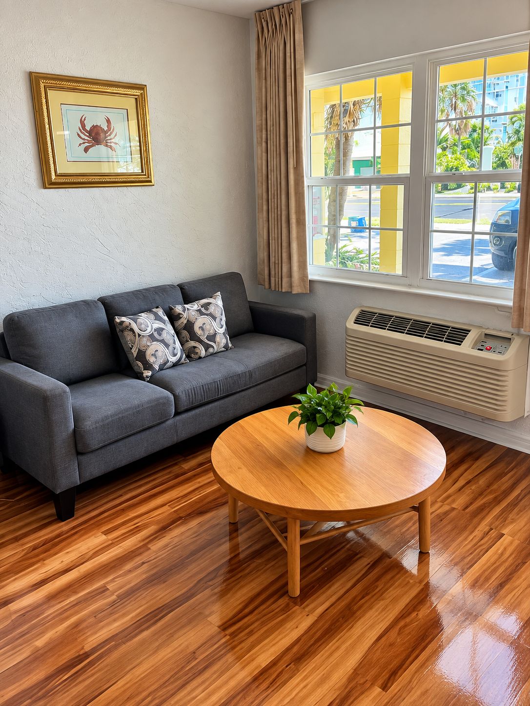 A room with a grey sofa, a round wooden table with a plant, a framed picture on the wall, and a window with curtains.