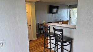 A compact kitchen with a bar area, a television mounted above the counter, and a door leading to another room. - Studio 1 Motel (Daytona Beach)