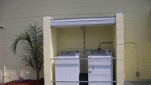 A laundry room with two stacked washers and dryers, a small plant, and a concrete floor. - Studio 1 Motel (Daytona Beach)
