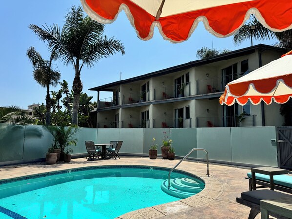 A swimming pool with a poolside area, including chairs and a table, surrounded by a building with balconies and potted plants.