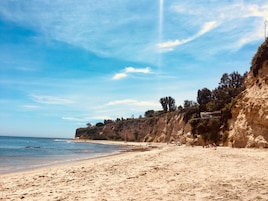 A beach with a sandy shore, a cliff with vegetation, and a clear sky.