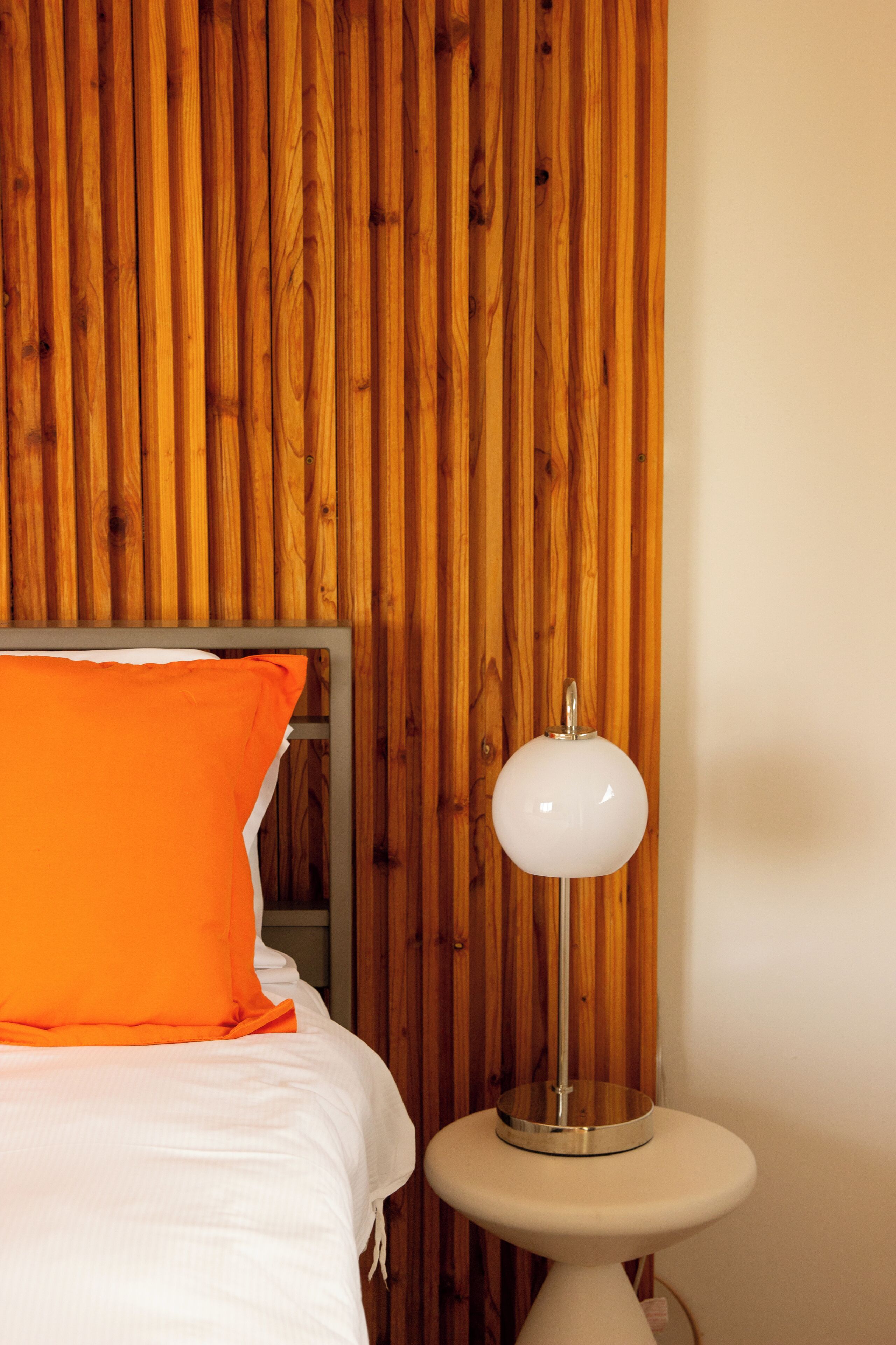 A room with a wooden slat wall, a bed with white bedding and an orange pillow, and a white table lamp.