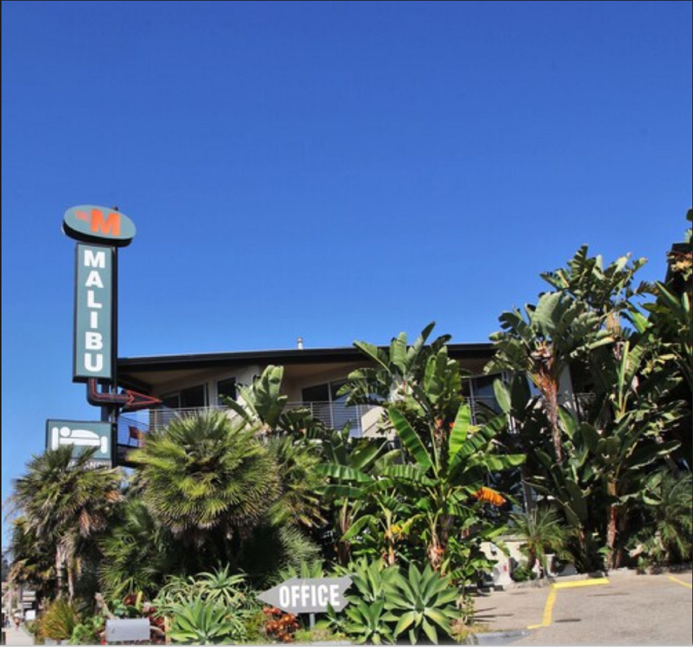 Un hotel con un grande cartello che recita 'MALIBU', circondato da rigogliosa vegetazione tropicale e un cielo azzurro limpido.