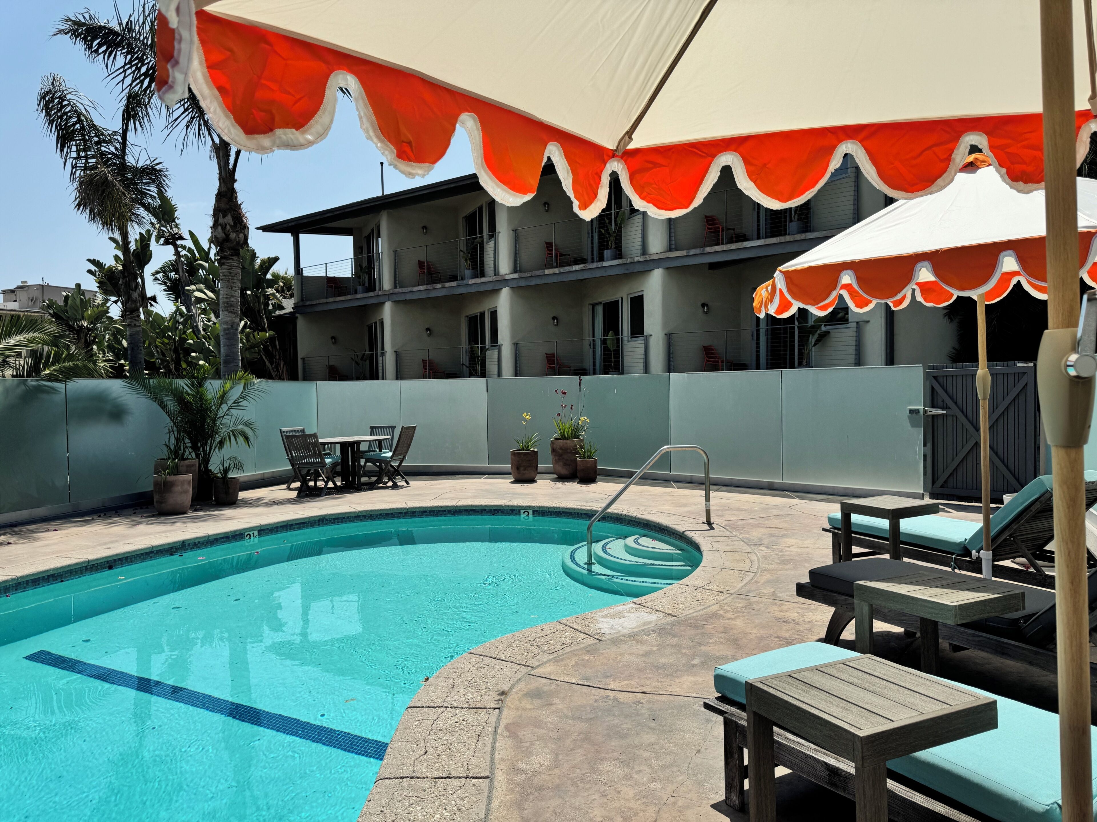 A swimming pool with lounge chairs and a table, surrounded by potted plants and a building with balconies.