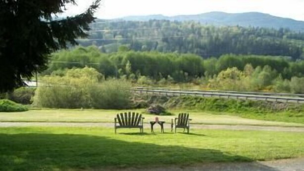 A grassy area with two Adirondack chairs and a picnic table, overlooking a forested landscape with a road in the background. - Valley View Motel (Port Townsend)