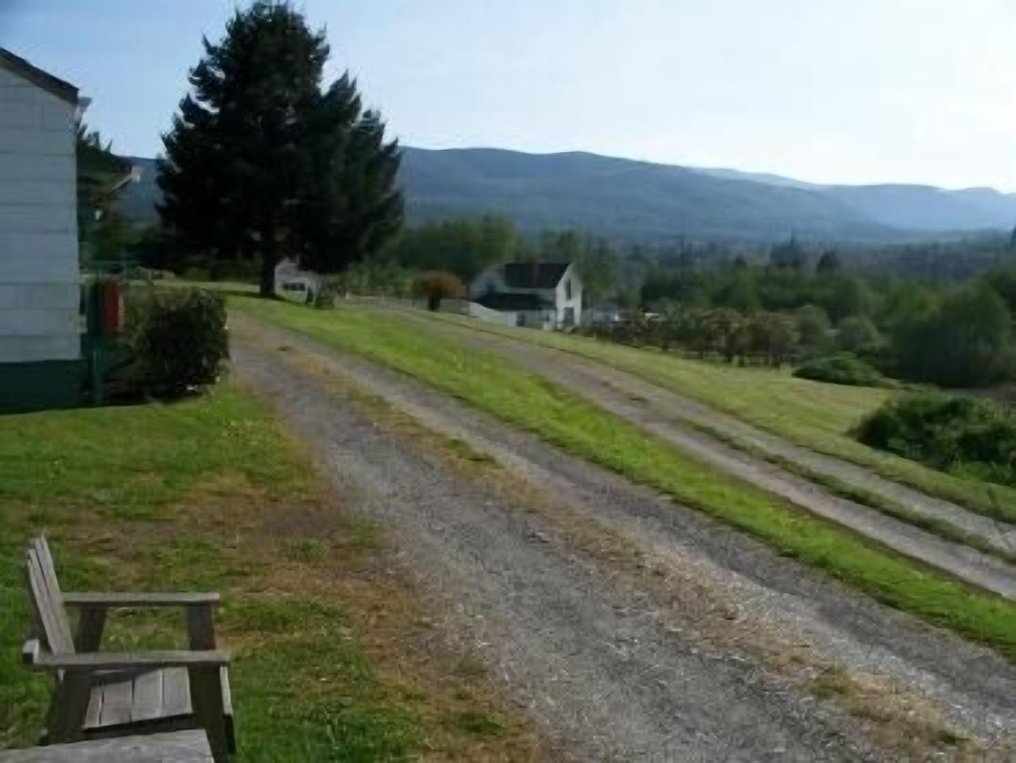 Entorno rural con un camino de grava que lleva a un edificio blanco, rodeado de vegetación y montañas al fondo.