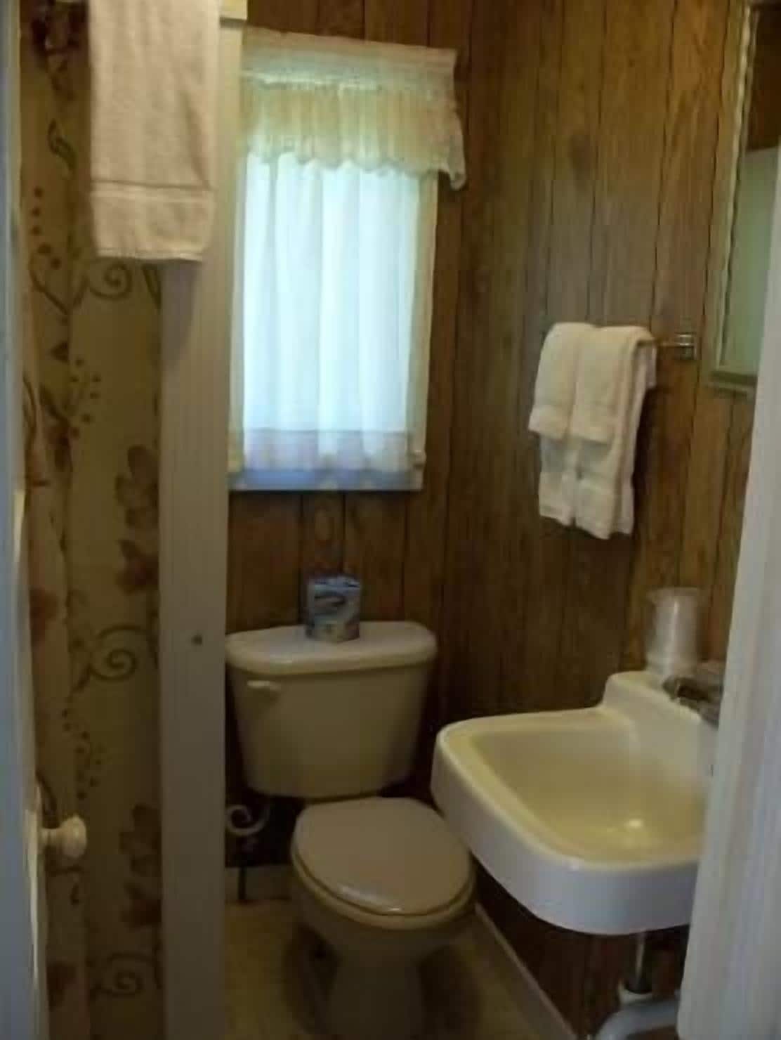 A small bathroom with wooden walls, a toilet, a sink, and a window with a curtain.
