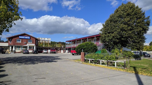 Un motel à l’extérieur rouge, avec une enseigne indiquant « Barrie Motel ». Plusieurs véhicules sont stationnés devant, et un grand arbre est visible sur la droite.