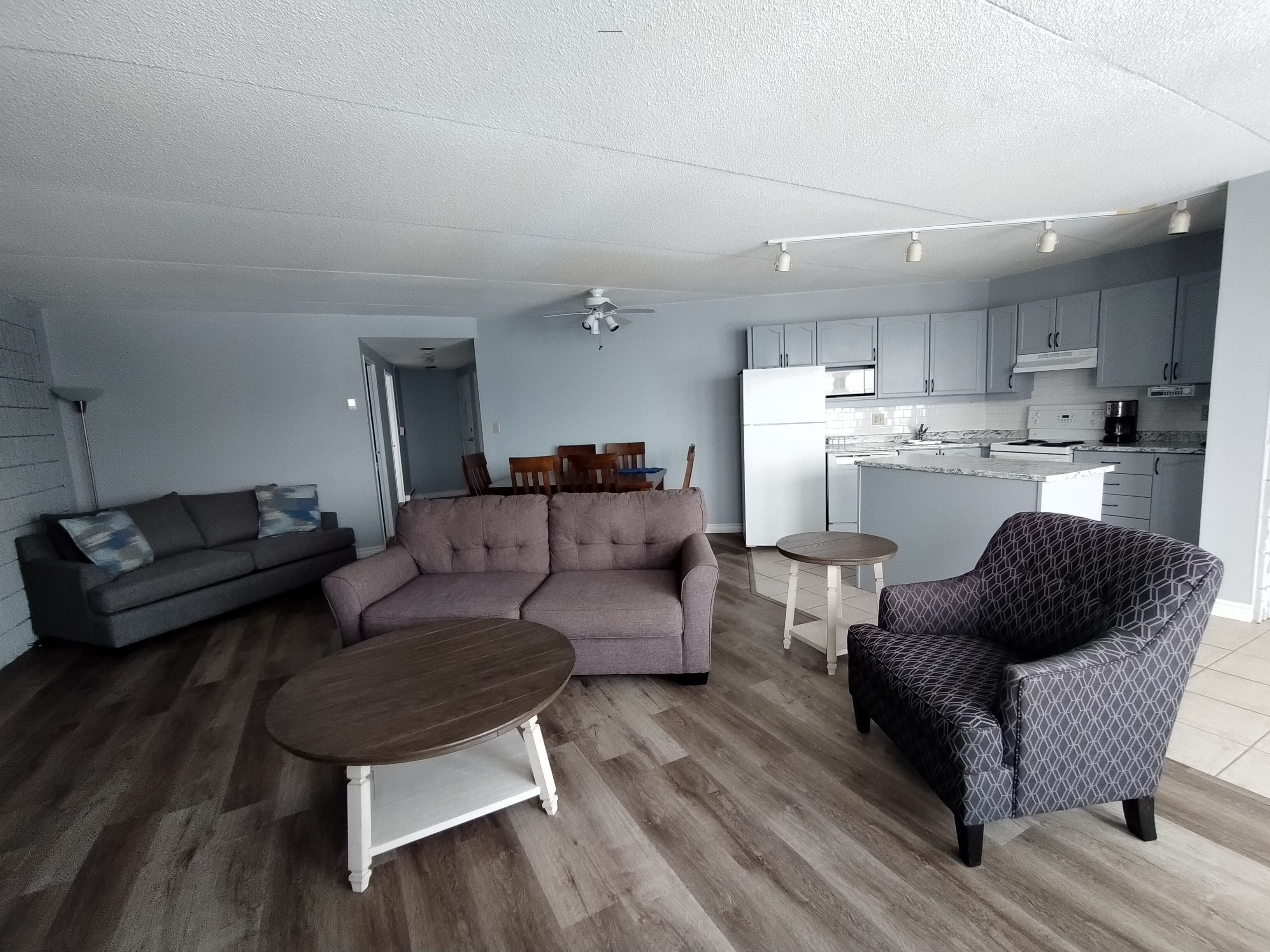 Un salon comprenant un canapé, une table basse ronde et un fauteuil à motifs. Il y a également un coin cuisine avec des placards et un réfrigérateur.