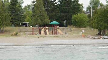 Playground area with slides and climbing structures, situated on a sandy surface, adjacent to a body of water.