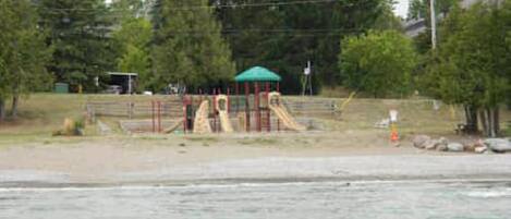 A playground with slides and a covered play structure, surrounded by trees and a body of water.